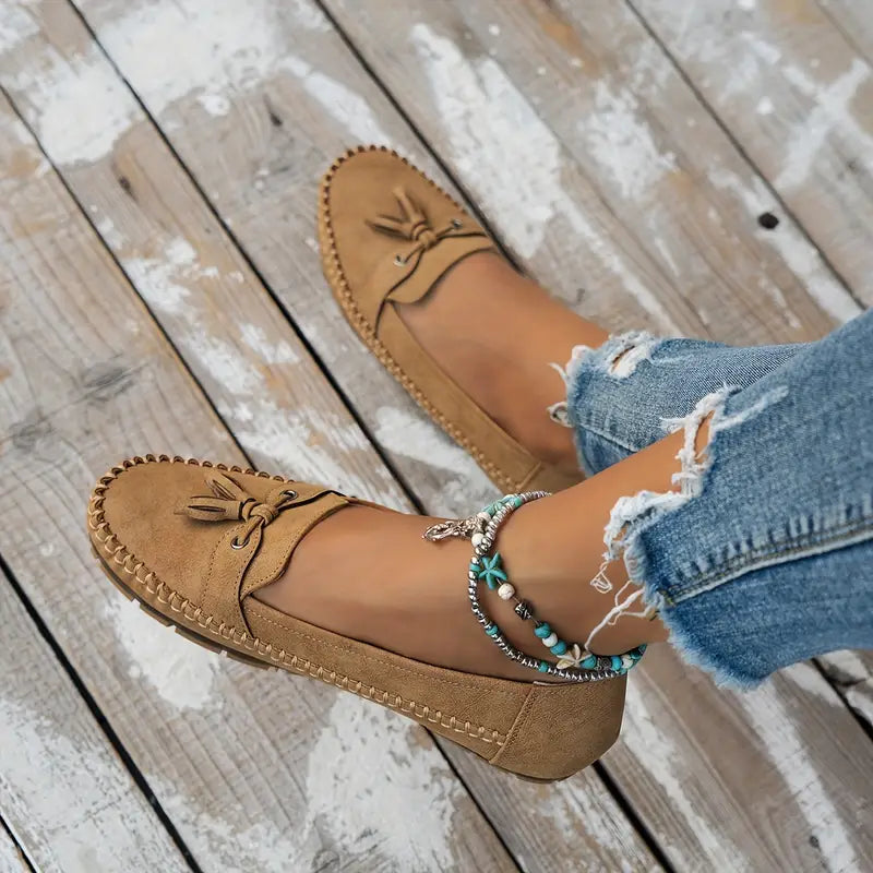 Des mocassins plats en suède beige avec un nœud décoratif à l'avant, associés à un jean déchiré et des bracelets bohèmes colorés. Ce modèle élégant et confortable est parfait pour un look décontracté et moderne, offrant à la fois style et praticité au quotidien.