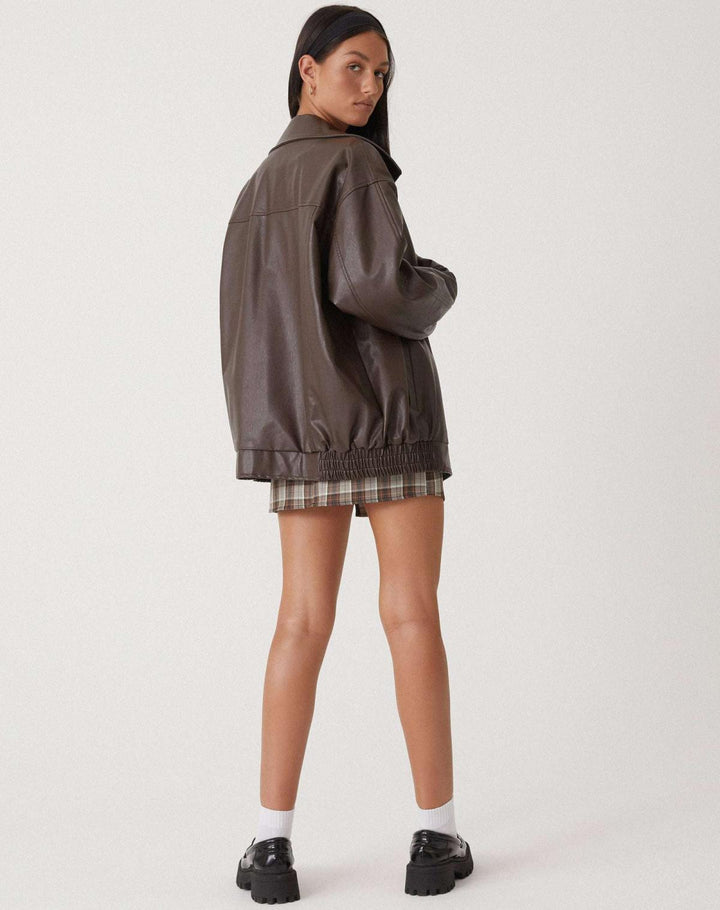 Vue arrière de la veste oversize en PU pour femme, modèle Cécile. Veste en simili-cuir marron avec coupe ample et taille élastiquée, portée avec une jupe à carreaux. Un style moderne et confortable pour l'hiver.