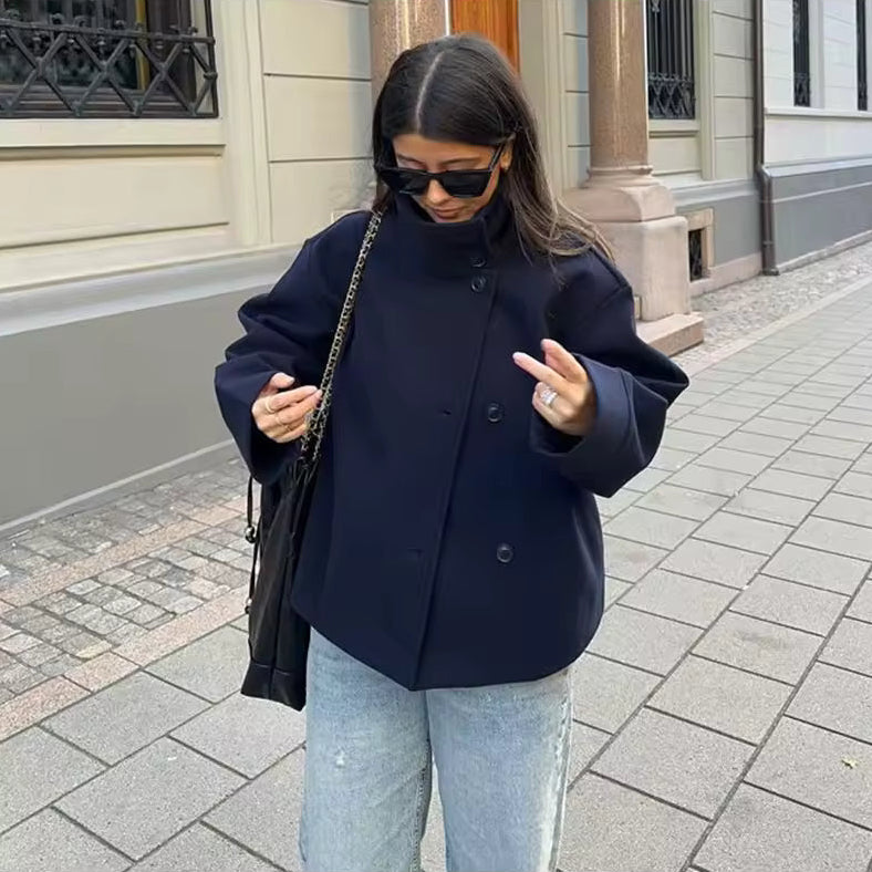 Femme marchant en ville portant une veste ample bleu marine à col montant et boutons sur le devant, associée à un jean clair et des lunettes de soleil noires. Elle tient un sac à bandoulière noire à chaîne métallique, créant un look moderne et décontracté idéal pour la mi-saison ou les journées fraîches d’été.