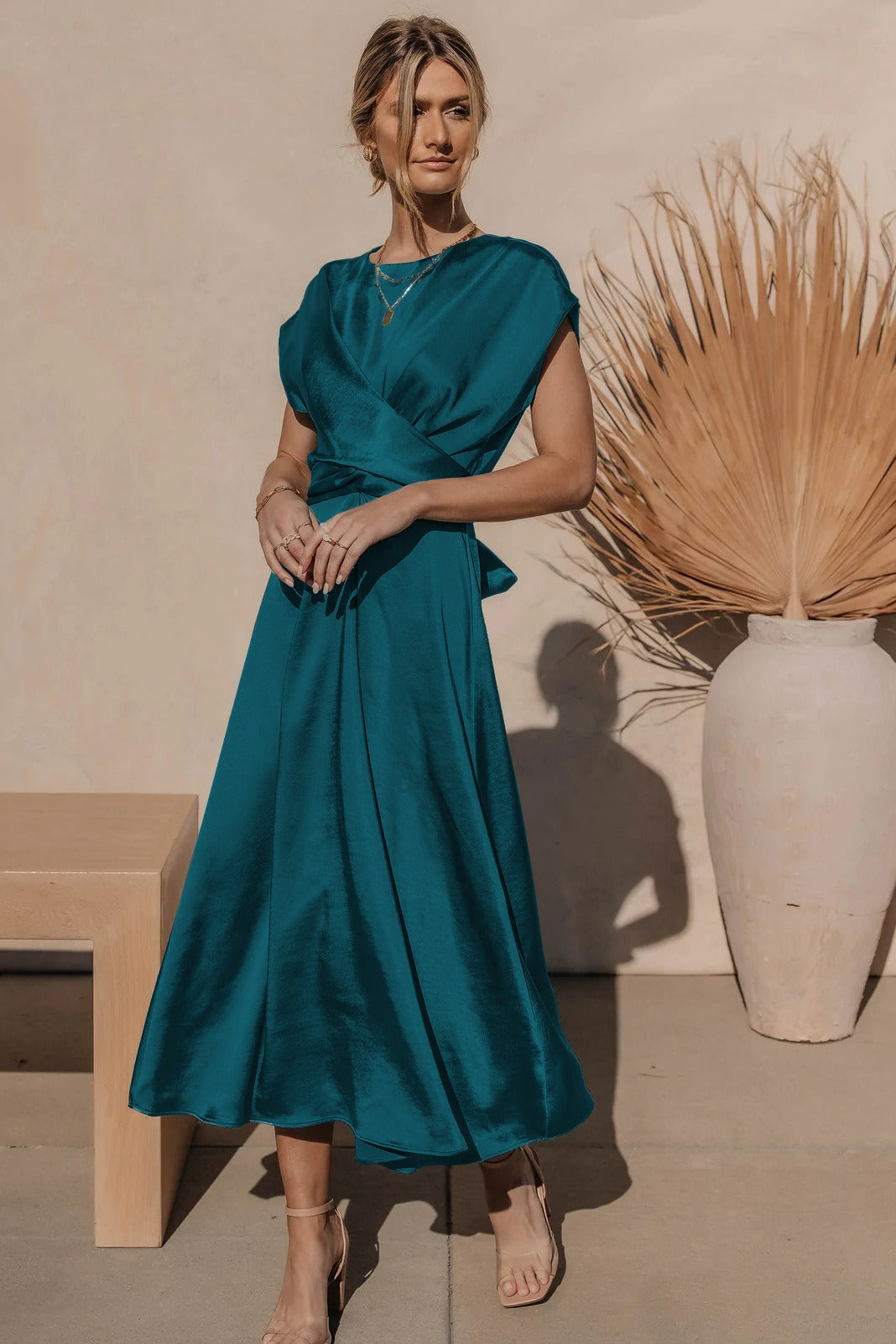 Femme posant de profil avec la robe fluide Chaïma bleu canard satinée, décolleté croisé et taille drapée, jupe midi évasée, portée avec sandales beige, devant un mur clair et un grand vase décoratif.