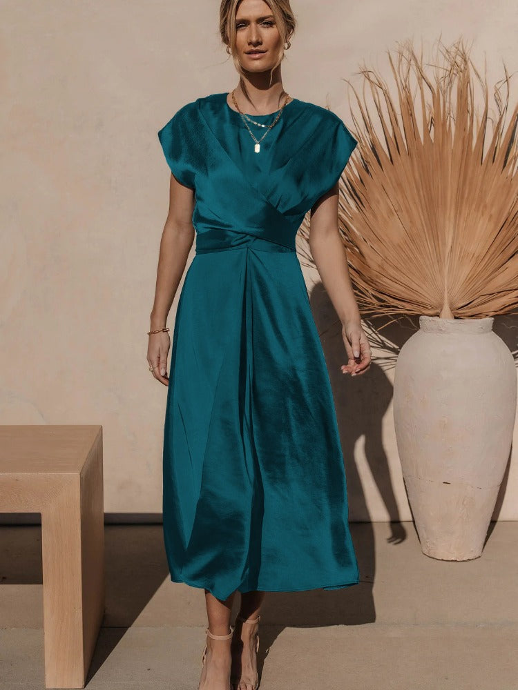 Femme portant la robe fluide Chaïma couleur bleu canard satinée, à décolleté croisé et taille drapée, longueur midi, avec sandales beige, devant un mur clair et un grand vase décoratif.