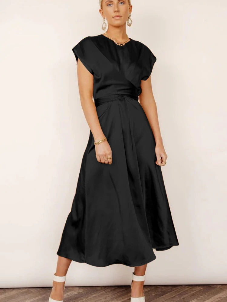 Femme portant la robe fluide Chaïma noire satinée, à décolleté croisé et taille drapée, longueur midi, avec bijoux dorés et sandales à brides, sur fond clair.