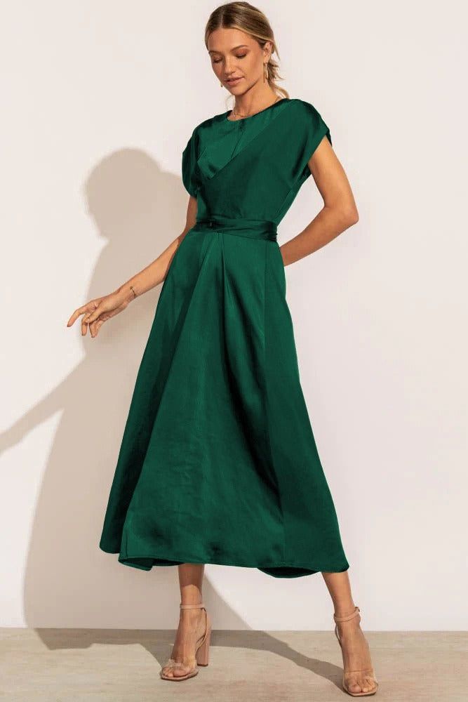 Femme portant la robe fluide Chaïma vert émeraude satinée, avec taille ceinturée et jupe midi évasée, posant sur fond clair avec sandales à talons nude.