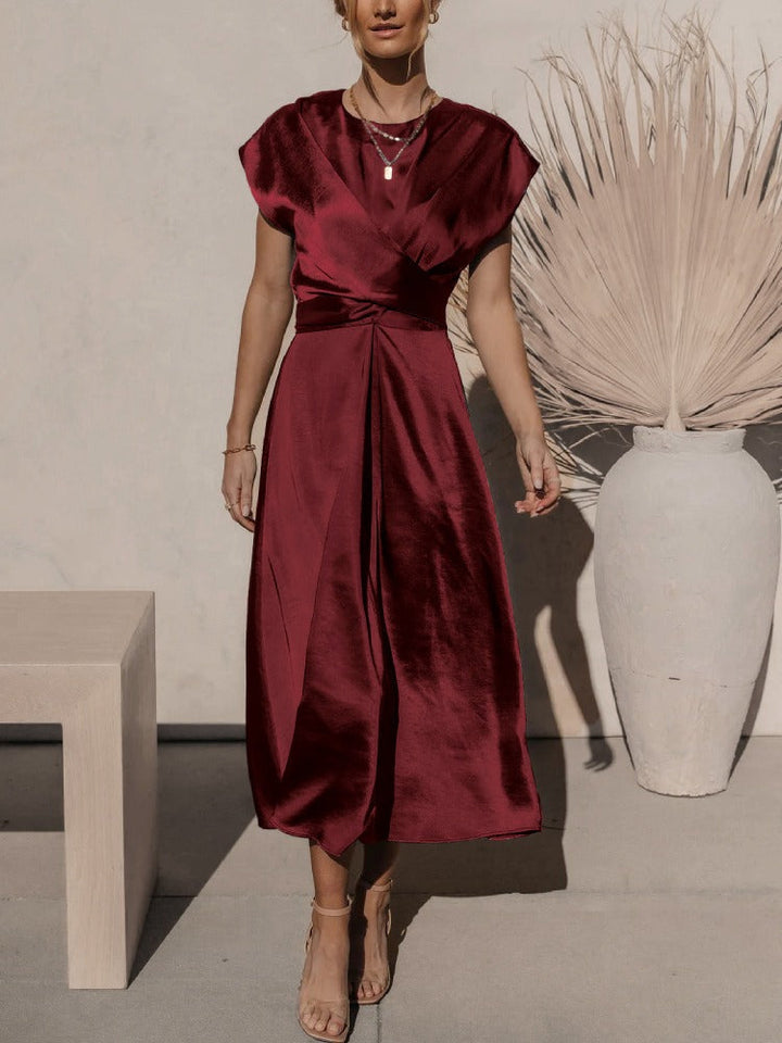 Femme portant la robe fluide Chaïma couleur bordeaux satinée, à décolleté croisé et taille drapée, longueur midi, avec sandales beige, devant un mur clair et un grand vase décoratif.
