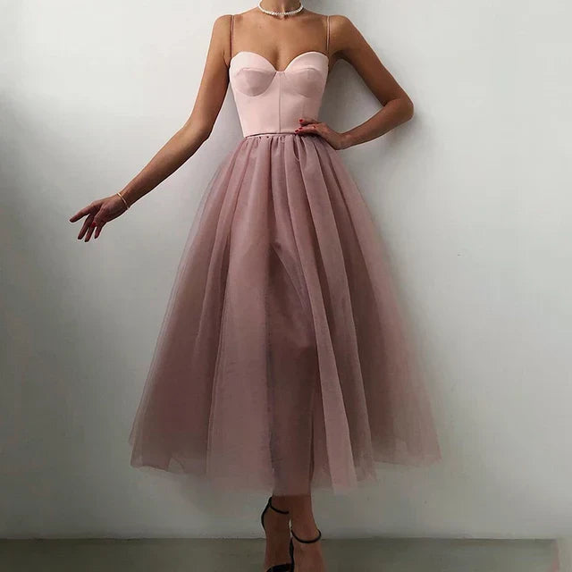 La femme porte une robe de soirée sans bretelles en tulle rose doux avec un décolleté en cœur. La robe présente une jupe volumineuse qui tombe élégamment jusqu'aux mollets, accompagnée d'un délicat collier ras du cou en perles. Elle pose gracieusement, mettant en valeur la silhouette féminine de la robe, idéale pour les événements formels.