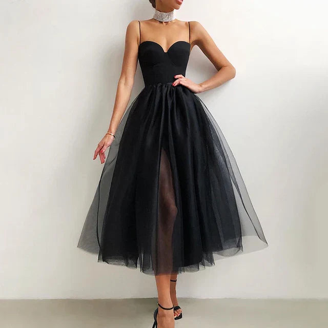 Une femme porte une robe de tulle noire sans bretelles avec un décolleté en cœur et une fente à l'avant. La robe présente une jupe volumineuse et une silhouette fluide, idéale pour les soirées élégantes. Elle accessoirise avec un collier ras du cou scintillant et des talons hauts, se tenant devant un fond neutre pour mettre en valeur le design gracieux de la robe.