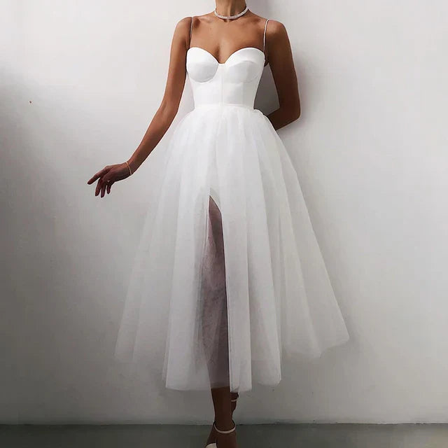 Une femme porte une robe de tulle blanche sans bretelles avec un décolleté en cœur, comportant une fente à l'avant. La robe présente une silhouette fluide et élégante, parfaite pour les événements formels en soirée. Elle accessoirise avec un délicat collier ras du cou et se tient devant un fond neutre, mettant en valeur le tissu léger et aérien de la robe.