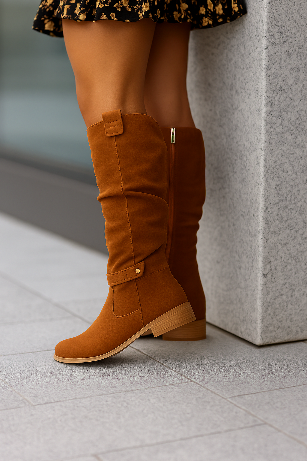 Bottes montantes en daim marron portées par une femme, montrées en gros plan alors qu’elle est appuyée contre un pilier en pierre. Les bottes présentent une tige souple légèrement plissée, une bride décorative à bouton au niveau de la cheville et un talon bas en bois, offrant un style moderne et chaleureux.