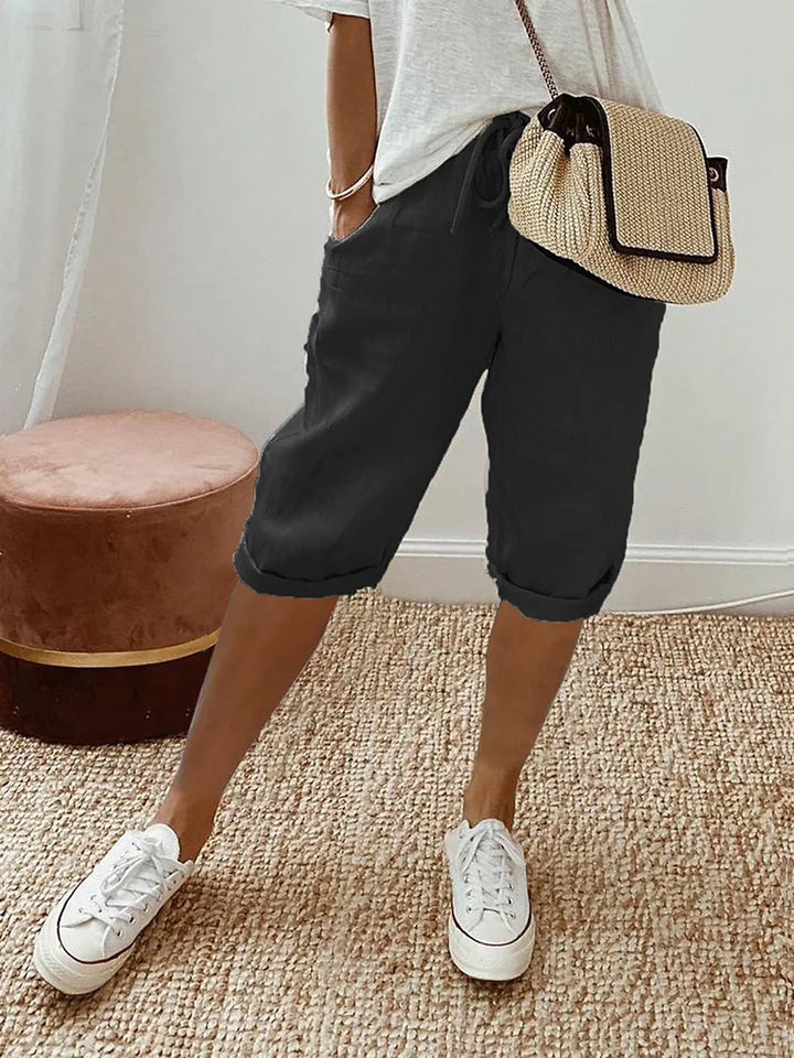 Femme portant un pantalon court noir à cordon, coupe décontractée, avec poches latérales, associé à un t-shirt clair, des baskets blanches et un petit sac bandoulière en paille, dans un intérieur lumineux au style naturel.