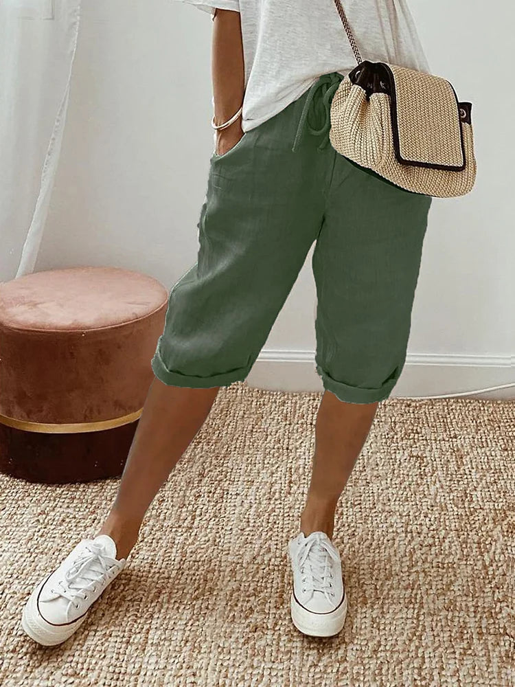 Femme portant un pantalon court vert à cordon, coupe décontractée avec poches latérales, associé à un t-shirt clair, des baskets blanches et un sac bandoulière en paille, dans un intérieur lumineux au style naturel.