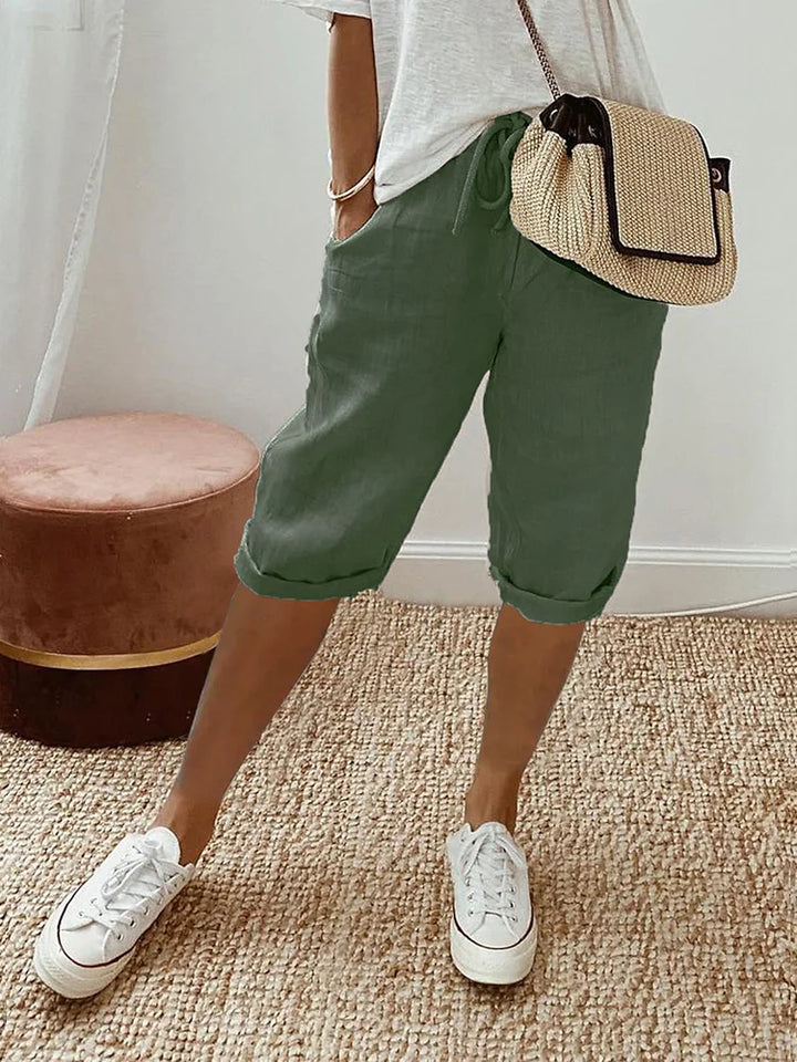 Femme portant un pantalon court vert à cordon, coupe décontractée avec poches latérales, associé à un t-shirt clair, des baskets blanches et un sac bandoulière en paille, dans un intérieur lumineux au style naturel.