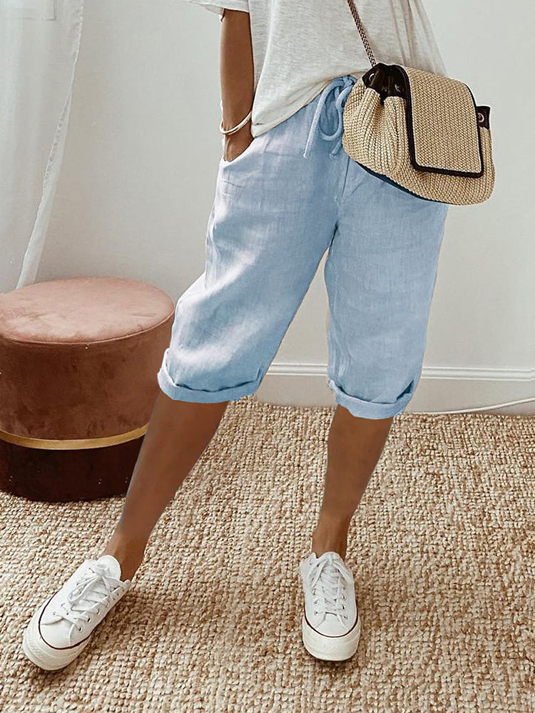 Femme portant un pantalon court bleu clair à cordon, coupe décontractée avec poches latérales, associé à un t-shirt clair, des baskets blanches et un sac bandoulière en paille, dans un intérieur lumineux au style naturel.