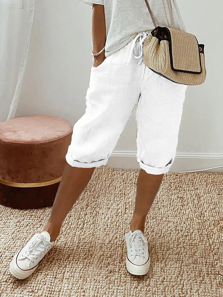 Femme portant un pantalon court blanc à cordon, coupe décontractée avec poches latérales, associé à un t-shirt clair, des baskets blanches et un sac bandoulière en paille, dans un intérieur lumineux au style naturel.