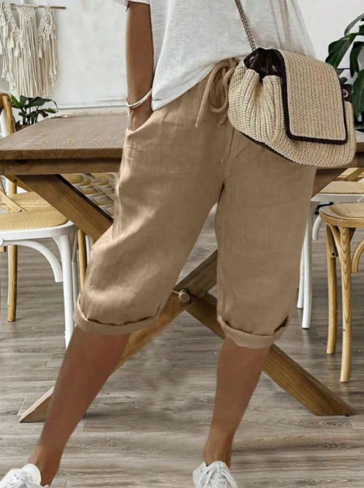 Femme portant un pantalon court beige à cordon, coupe décontractée avec poches latérales, associé à un t-shirt clair, des baskets blanches et un sac bandoulière en paille, dans un intérieur chaleureux au style naturel.