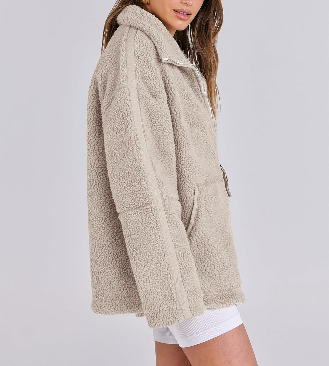 Veste en tissu peluche beige clair pour femme, vue de profil. Modèle à la texture douce et moelleuse, avec col montant, fermeture éclair à l’avant et poche latérale visible. Sa coupe ample et confortable apporte un style moderne et décontracté, parfait pour les journées froides d’automne et d’hiver.