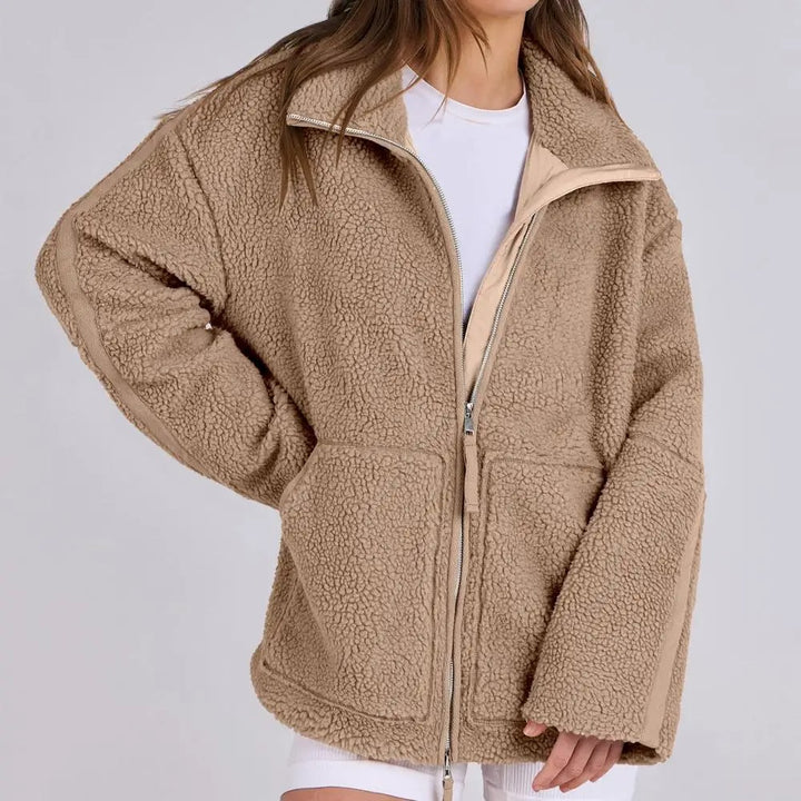 Veste en peluche marron clair pour femme, douce et moelleuse. Ce modèle est doté d'une fermeture éclair argentée sur le devant, d'un col montant et de deux grandes poches plaquées. Sa coupe ample et son design moderne lui confèrent un style à la fois décontracté et confortable, idéal pour les fraîches journées d'automne et d'hiver.