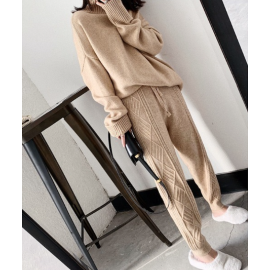 Une femme porte un ensemble confortable composé d'un pull beige à col haut et d'un pantalon de jogging beige à motif en tricot avec des jambes légèrement resserrées. Elle porte des chaussons blancs et tient un petit sac à main noir, créant ainsi un look décontracté et cosy.