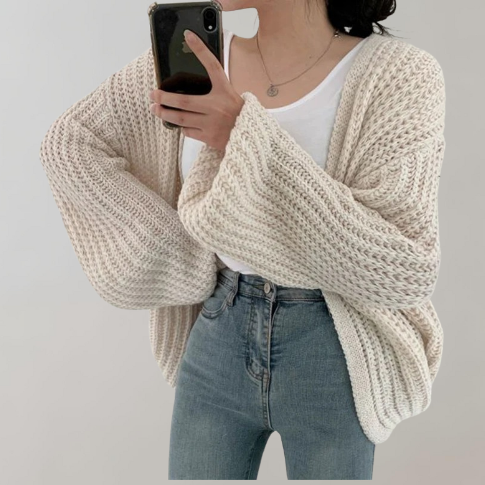 Une femme portant un cardigan blanc oversize en maille à manches longues et volumineuses, associé à un débardeur blanc et un jean taille haute, se prend en photo devant un fond neutre.