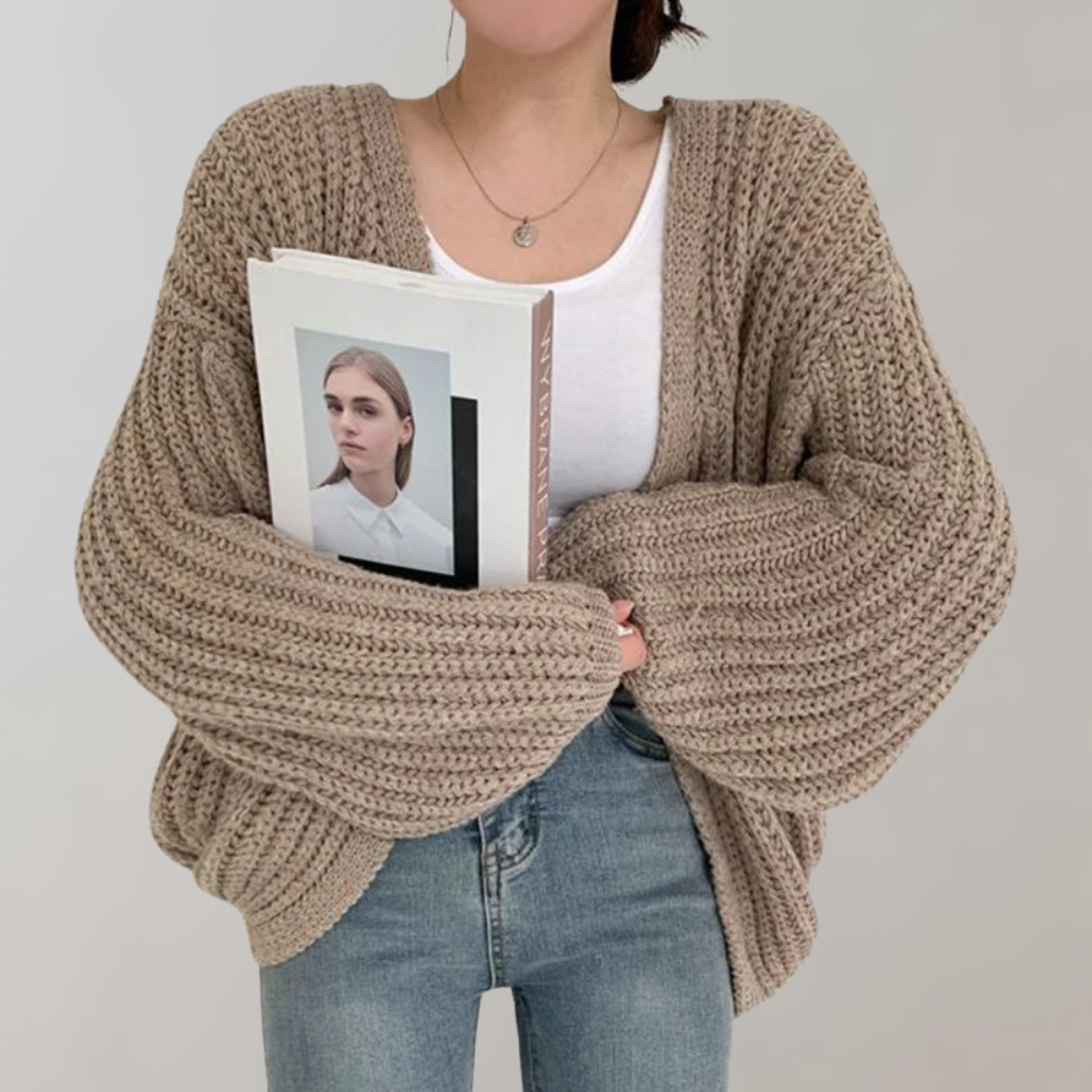 Une femme portant un cardigan oversize en maille épaisse de couleur kaki, à manches longues et volumineuses, associé à un débardeur blanc et un jean taille haute, tient un livre contre elle devant un fond neutre.