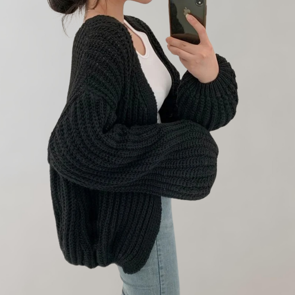 Femme portant un cardigan oversize en maille noire, à manches longues et volumineuses, associé à un débardeur blanc et un jean, se prenant en photo devant un fond neutre.