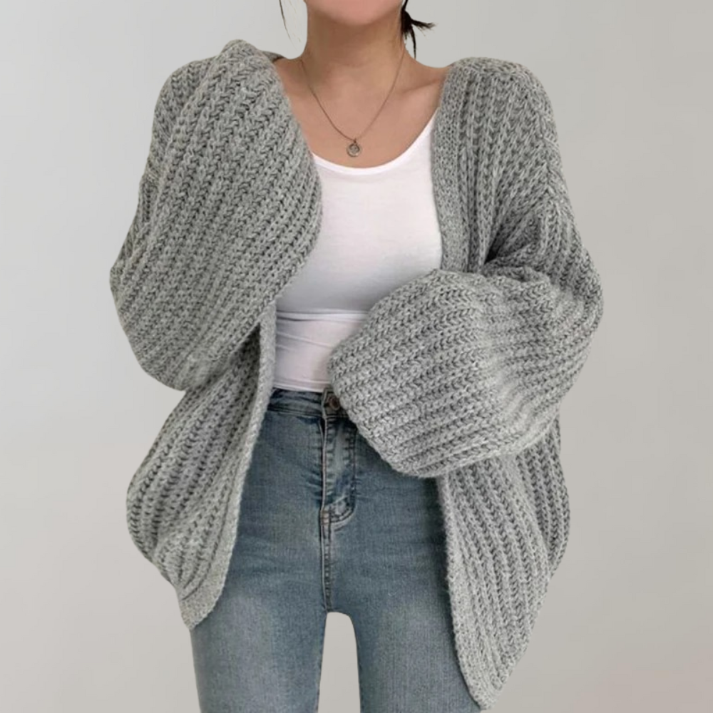 Femme portant un cardigan gris en maille oversize à manches larges et volumineuses, associé à un débardeur blanc et un jean taille haute, photographiée sur un fond neutre.
