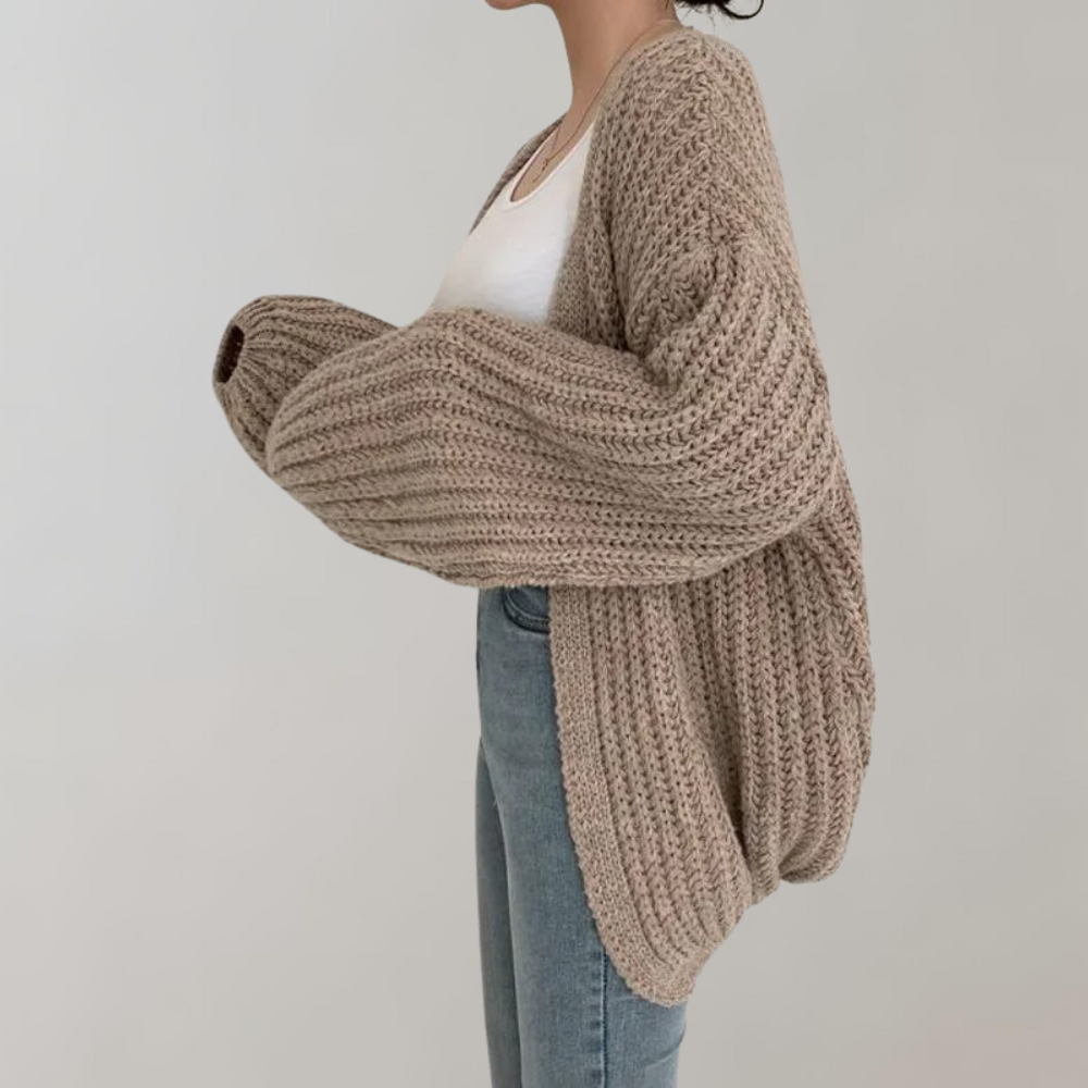 Femme vue de profil portant un cardigan oversize kaki en grosse maille à manches larges et volumineuses, associé à un débardeur blanc et un jean taille haute, photographiée devant un fond neutre.