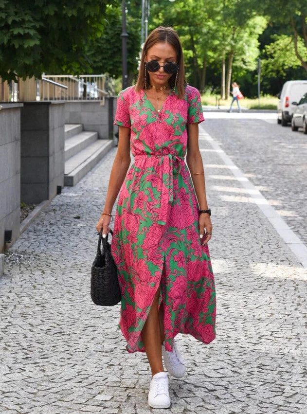 Robe midi femme Demi au design moderne avec col en V et imprimé floral rose et vert. Coupe fluide avec taille cintrée et lien à nouer, idéale pour un look estival élégant et décontracté, portée avec des baskets blanches et un sac noir.