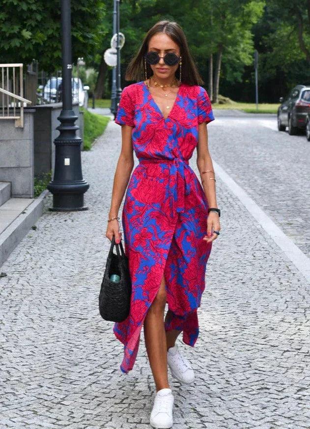 Robe midi femme Demi au style moderne avec col en V et imprimé floral rouge et bleu. Coupe fluide avec taille cintrée et lien à nouer, portée avec des baskets blanches et un sac noir pour un look estival élégant et décontracté.