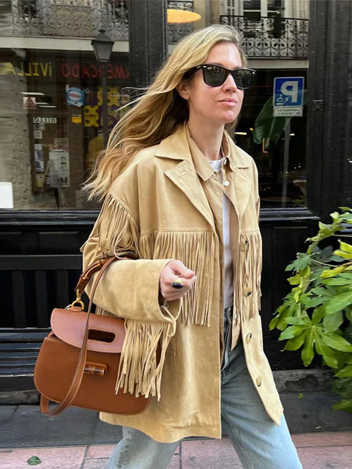 Veste à franges pour femme, modèle Domitille. Veste beige en suède avec franges sur les épaules et les manches, portée avec une chemise et un sac à main en cuir. Un look tendance et décontracté pour un style chic et moderne.