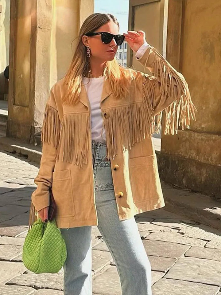 Veste à franges pour femme, modèle Domitille. Veste beige en suède avec franges sur les manches et le devant, portée avec un t-shirt blanc et un jean taille haute. Complétée par un sac vert vif, un look tendance et dynamique pour un style moderne.
