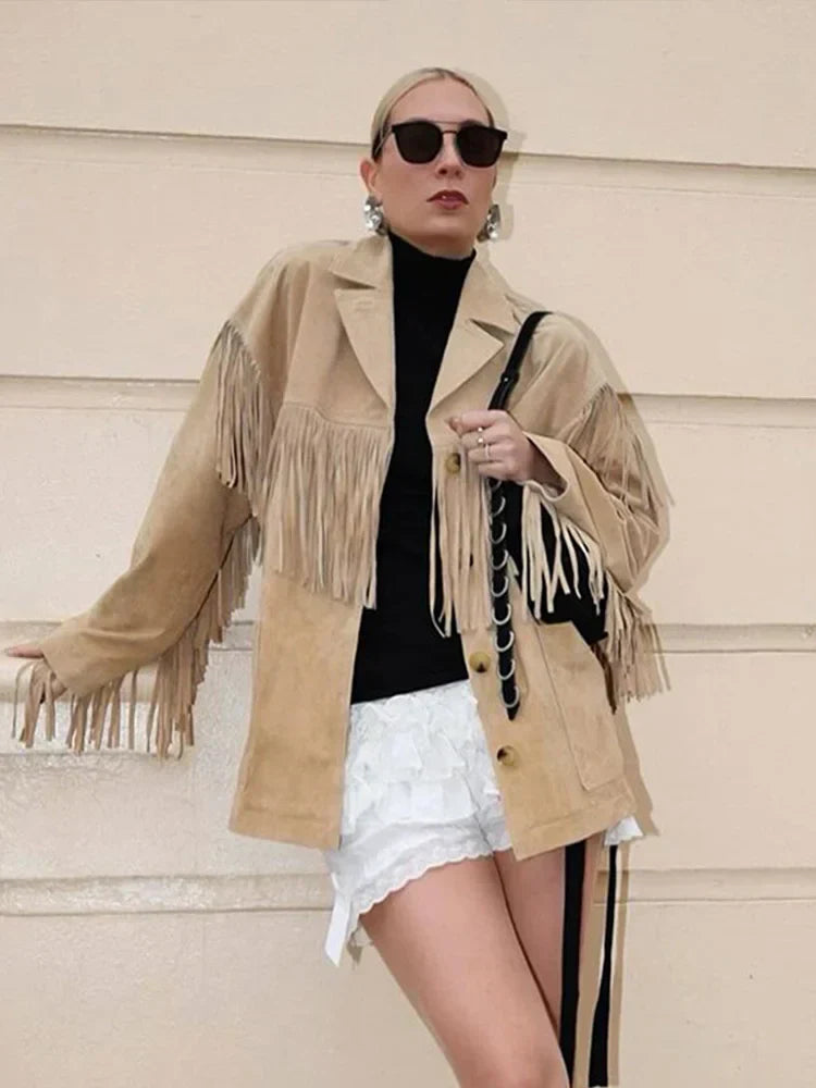 Veste à franges pour femme, modèle Domitille. Veste en suède beige avec franges sur les manches et le bas, portée avec un t-shirt noir et des shorts blancs. Accessoirisée avec un sac à main élégant, un look tendance et moderne.