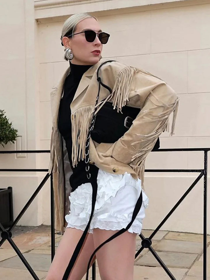 Veste à franges pour femme, modèle Domitille. Veste beige en suède avec franges sur les manches et le devant, portée avec un t-shirt noir, des shorts blancs et un sac noir élégant. Un look tendance et audacieux pour un style moderne et chic.