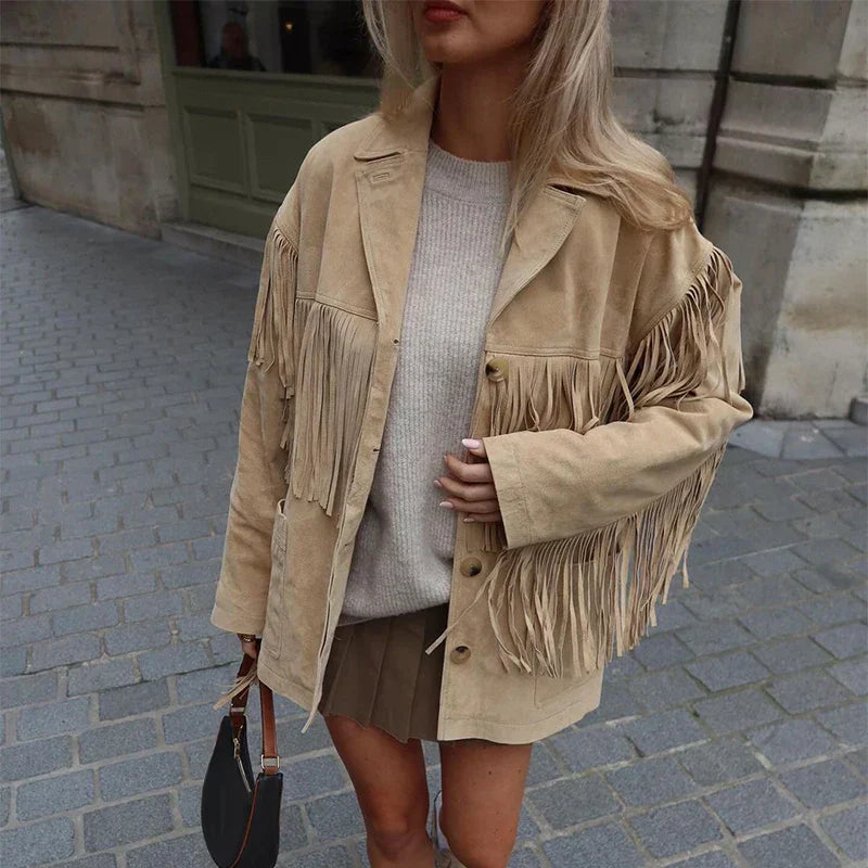 Veste à franges pour femme, modèle Domitille. Veste en suède beige avec franges sur les épaules et les manches, portée avec un pull beige et une jupe courte. Accessoirisée avec un sac à main, un look tendance et élégant, idéal pour une tenue décontractée et stylée.