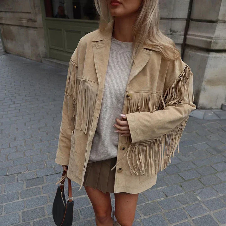 Veste à franges pour femme, modèle Domitille. Veste en suède beige avec franges sur les épaules et les manches, portée avec un pull beige et une jupe courte. Accessoirisée avec un sac à main, un look tendance et élégant, idéal pour une tenue décontractée et stylée.