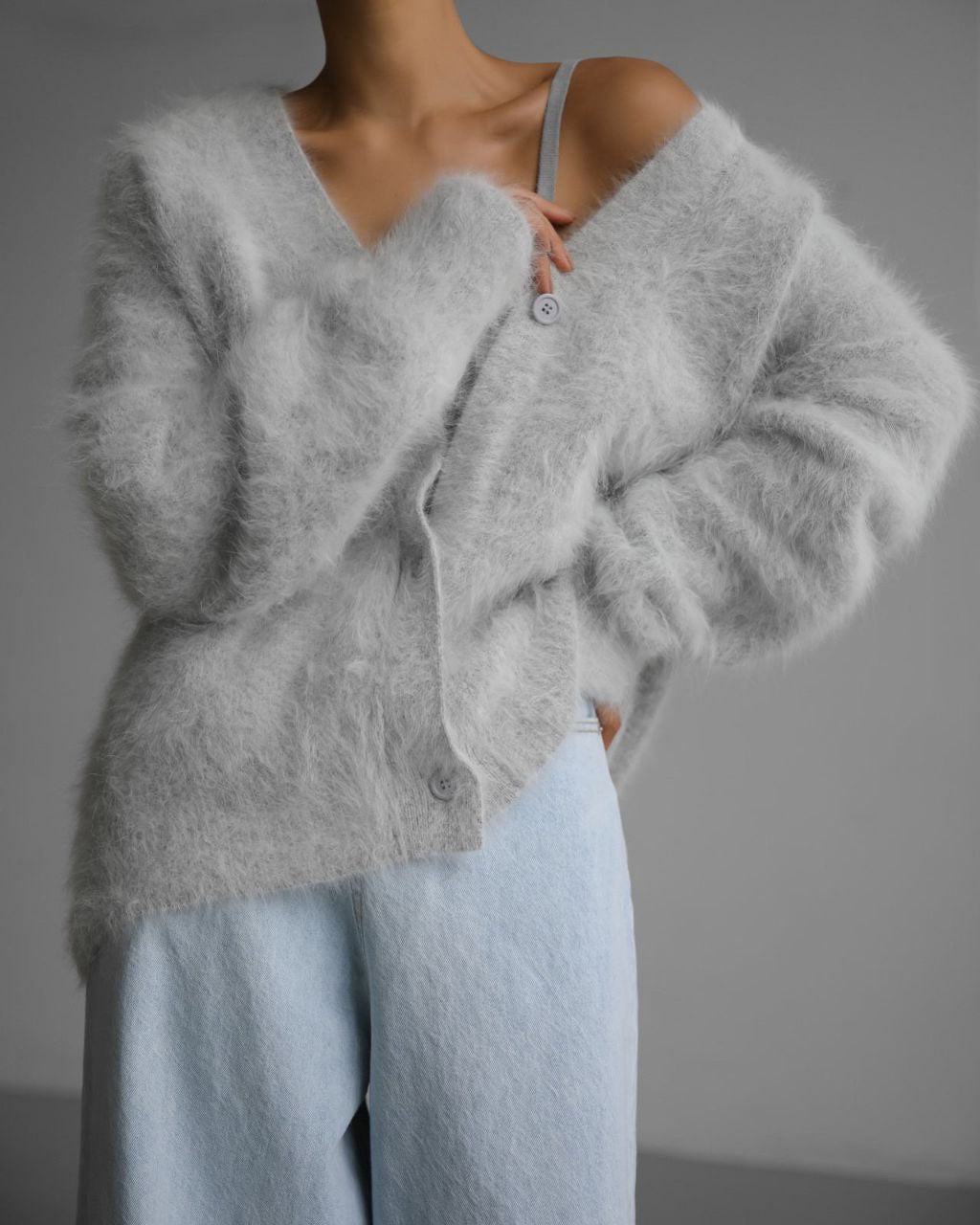 Un cardigan en laine douce de couleur grise, avec une coupe décontractée et des boutons au centre. Il est porté sur un haut léger, apportant une touche moderne et cosy à la tenue.