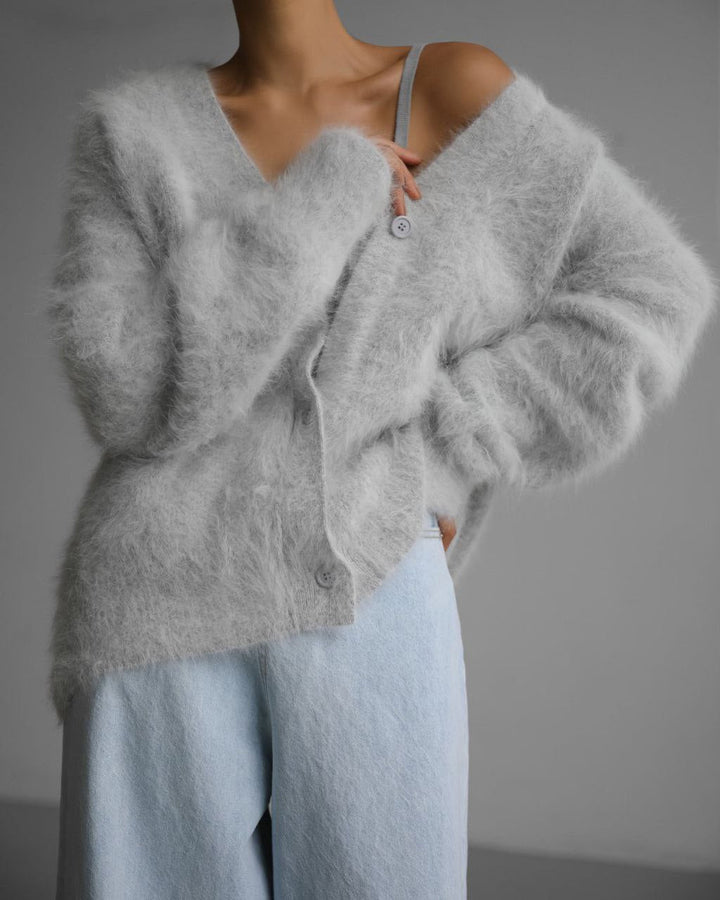 Un cardigan en laine douce de couleur grise, avec une coupe décontractée et des boutons au centre. Il est porté sur un haut léger, apportant une touche moderne et cosy à la tenue.