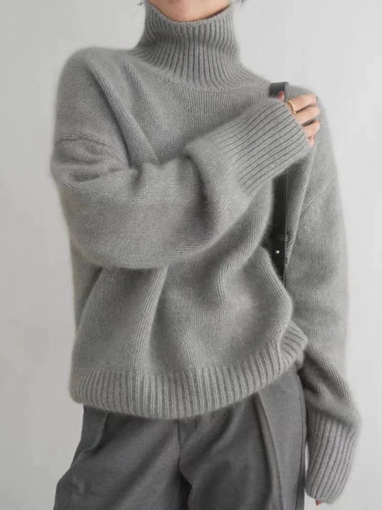 Pull à col roulé femme Éléonore gris en maille épaisse et douce, coupe ample avec col montant et larges bords côtelés aux poignets et à l’ourlet, porté avec un pantalon gris — idéal pour l’hiver.