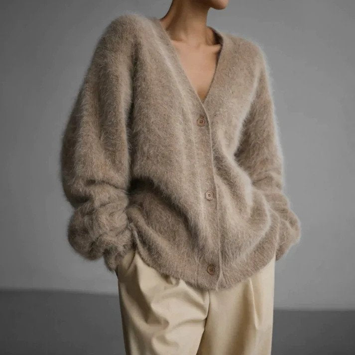 Femme portant un cardigan en maille duveteuse couleur beige, avec une coupe ample et un décolleté en V, fermé par des boutons en bois et associé à un pantalon large crème, photographiée sur un fond gris neutre.
