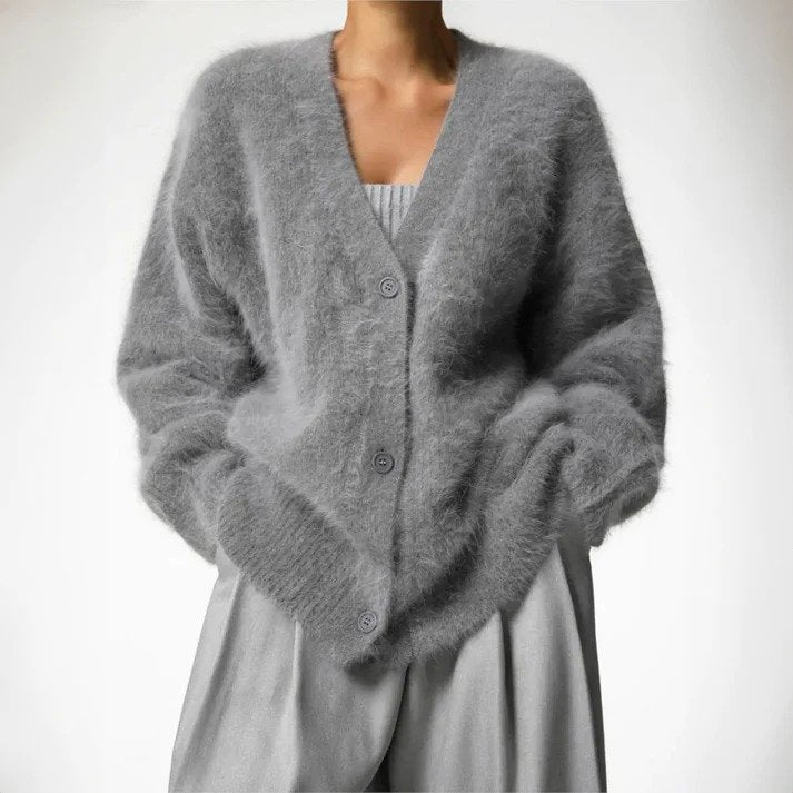 Femme portant un cardigan long en maille duveteuse gris, à coupe ample et décolleté en V, fermé par des boutons, associé à un haut plissé et un pantalon large assorti, photographiée sur un fond clair et minimaliste.