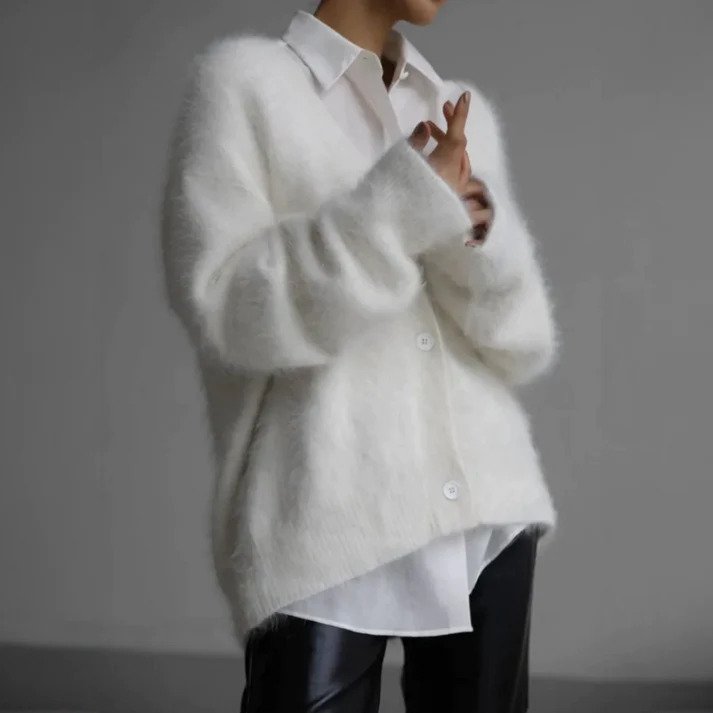 Femme portant un cardigan long en maille duveteuse blanc, à coupe ample et boutonné sur le devant, associé à une chemise blanche et un pantalon foncé, photographiée sur un fond gris neutre.
