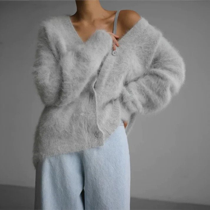 Femme portant un cardigan long en maille duveteuse gris clair, légèrement retombant sur une épaule, avec un décolleté en V et une coupe ample, associé à un pantalon large bleu pâle, photographiée sur un fond gris minimaliste.