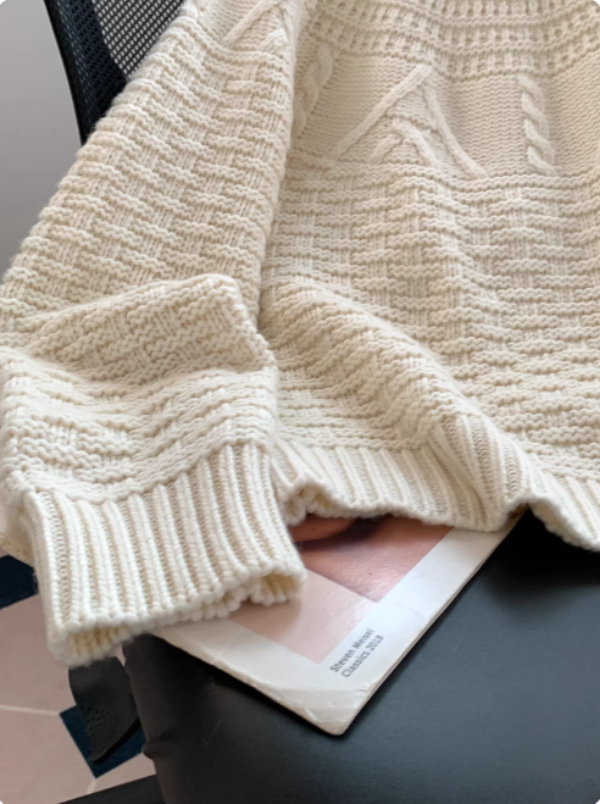 Gros plan sur un pull blanc en maille, montrant ses poignets côtelés et ses motifs texturés, posé sur une chaise avec un magazine en dessous.