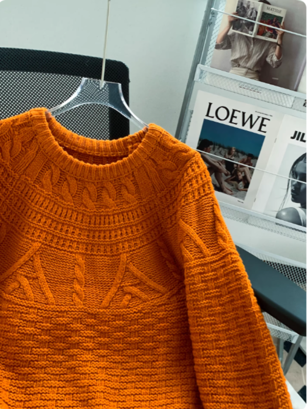 Pull en maille orange vif avec col rond et motifs texturés, présenté sur un cintre transparent dans un intérieur moderne avec un porte-magazines en arrière-plan.