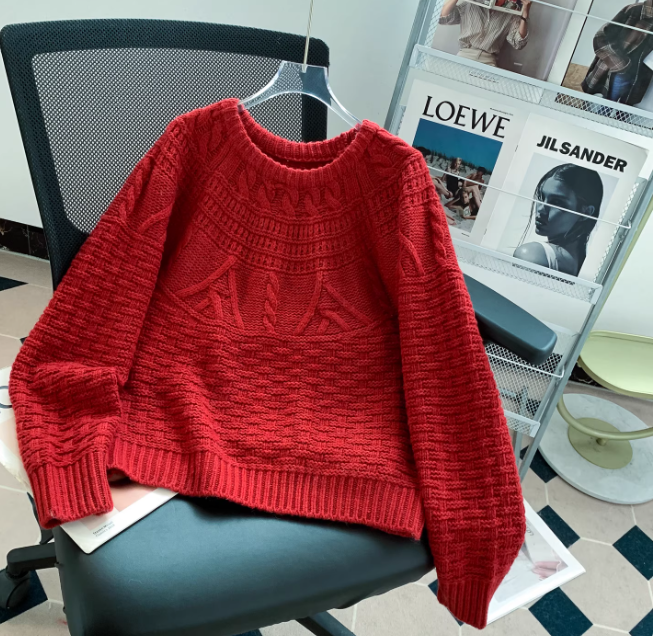 Pull en maille rouge à col rond, doté de motifs texturés et de finitions côtelées, posé sur une chaise de bureau avec un porte-magazines en arrière-plan dans un intérieur moderne.