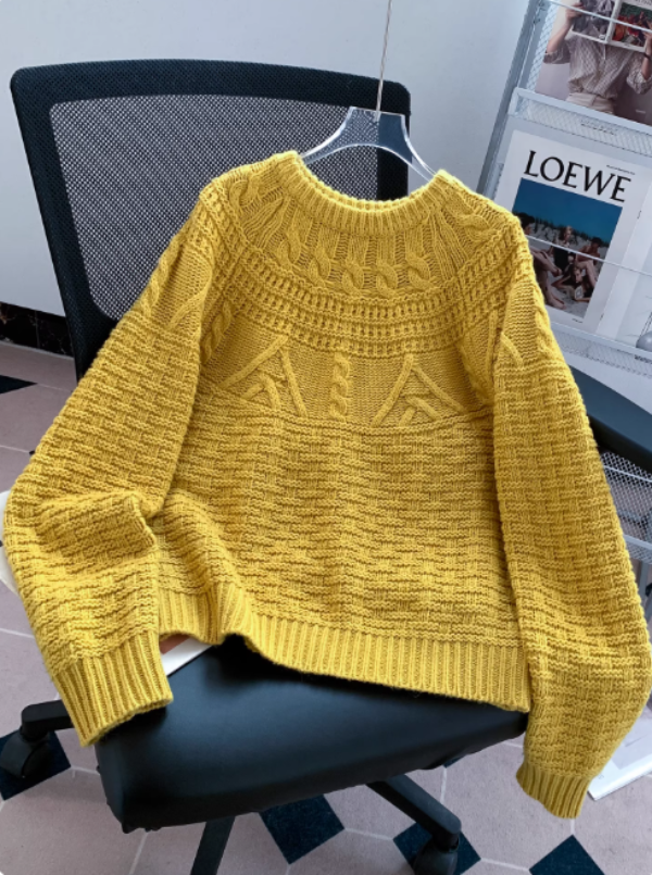 Pull en maille jaune moutarde avec col rond, motifs texturés et finitions côtelées, posé sur une chaise de bureau dans un intérieur moderne avec un porte-magazines en arrière-plan.