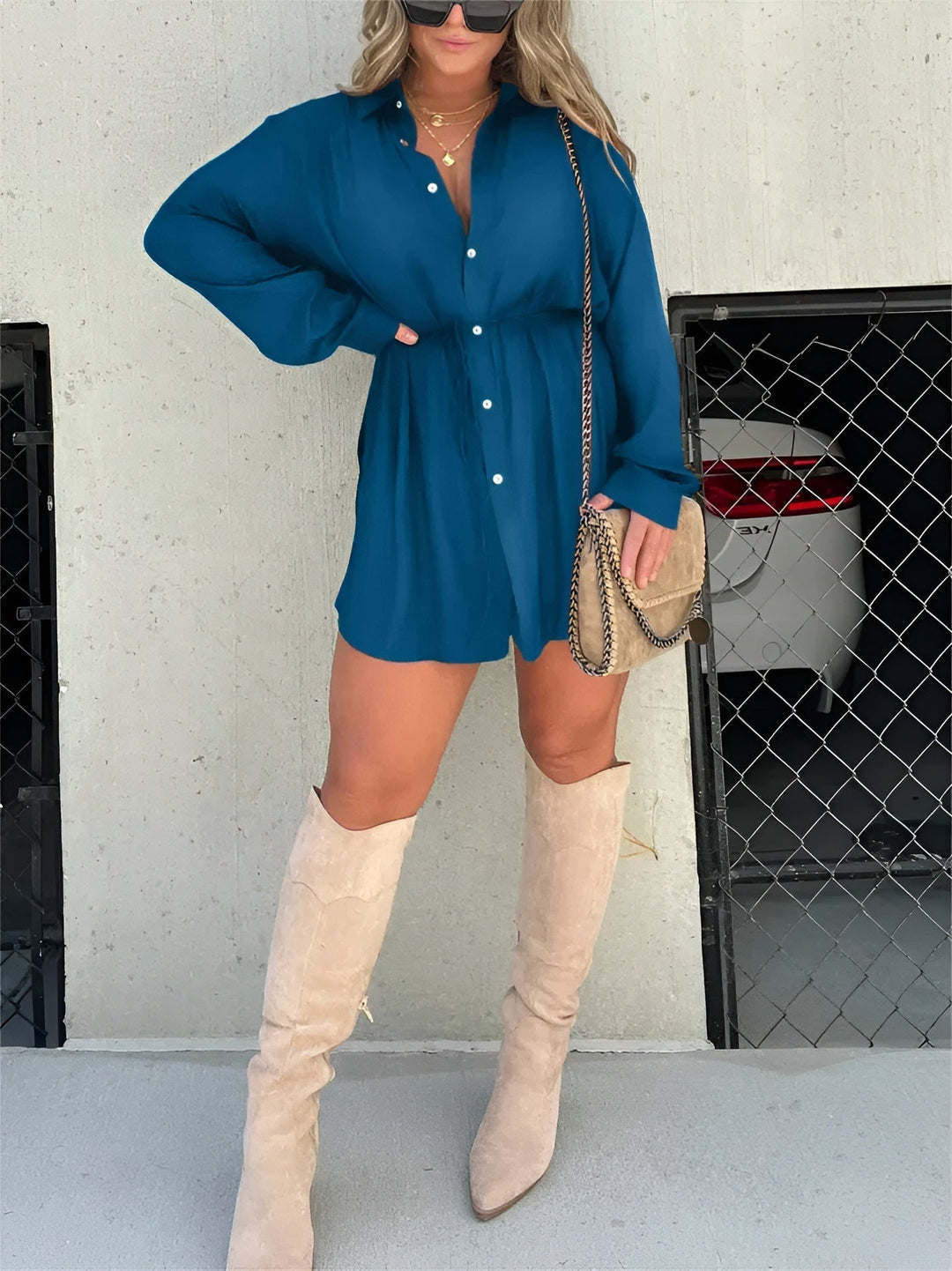 Cette chemise fluide d'un bleu profond, avec des boutons et une ceinture à nouer à la taille, crée un look chic et décontracté. Elle est portée avec des bottes hautes beiges et un sac à bandoulière, parfait pour un style moderne et élégant.
