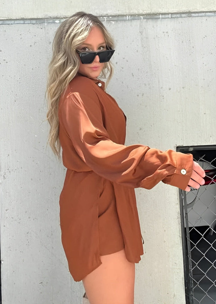 Cette chemise fluide de couleur marron terracotta, avec des boutons et une ceinture à nouer à la taille, crée une silhouette élégante et décontractée. Elle est portée avec des lunettes de soleil et un look moderne, parfait pour un style estival et chic.