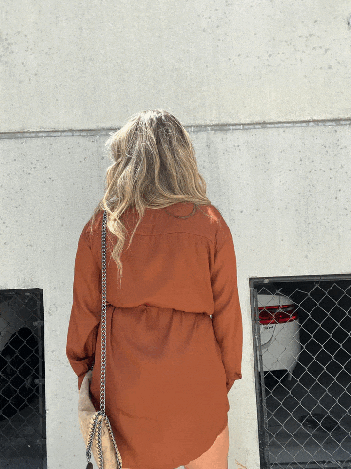 Cette chemise fluide de couleur marron terracotta, vue de dos, présente une coupe élégante et une ceinture à nouer à la taille, offrant une silhouette moderne et décontractée. Elle est portée avec un sac à bandoulière, parfaite pour un look chic et estival.