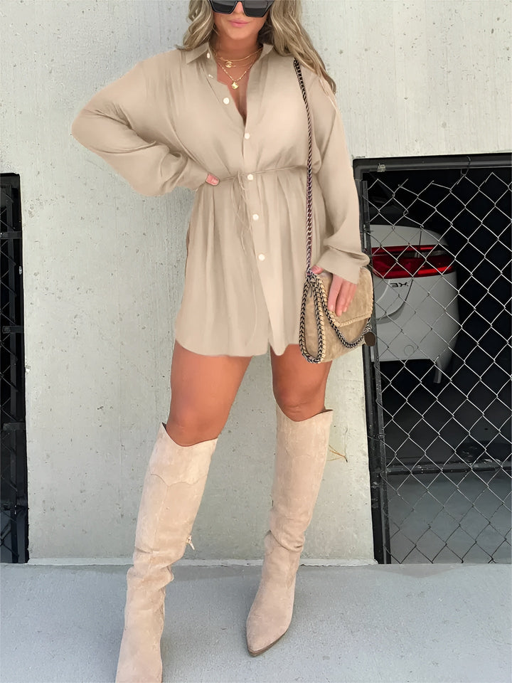 Chemise à manches longues pour femme, modèle tendance. Cette chemise fluide de couleur beige, avec des boutons et une ceinture à nouer à la taille, crée une silhouette élégante et décontractée. Elle est portée avec des bottes hautes beiges et un sac à bandoulière, parfait pour un look moderne et chic.