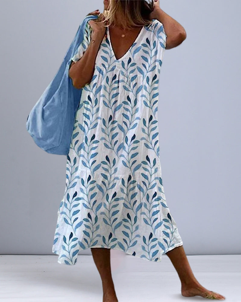Robe col V Elysia pour femme, coupe ample tendance avec imprimé feuilles bleues sur fond blanc, portée pieds nus avec un grand sac bleu, look estival décontracté.